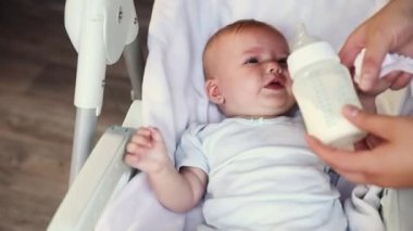 Şişeden süt içen ve beyaz arka planda kameraya bakan tatlı küçük bir kız. Bebek emzirme sütü yiyor ve evdeki beslenme sandalyesine uzanıyor. Annelik mutlu çocuk