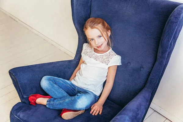 ジーンズの甘い女の子と白のTシャツは 屋内の自宅で白い明るいリビングルームでリラックス現代的な居心地の良い青い椅子に座っています 子供の頃の若者のリラックスコンセプト — ストック写真