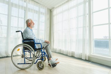Tekerlekli sandalyede oturan üzgün yaşlı adam huzurevinin penceresinden dışarı bakıyor. Engelli insanlar için tekerlekli sandalyedeki yaşlı felçli adam depresyonda. Yalnız yaşlı hasta adam tekerlekli sandalyede oturuyor.