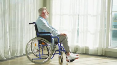 Tekerlekli sandalyede oturan üzgün yaşlı adam huzurevinin penceresinden dışarı bakıyor. Engelli insanlar için tekerlekli sandalyedeki yaşlı felçli adam depresyonda. Yalnız yaşlı hasta adam tekerlekli sandalyede oturuyor.