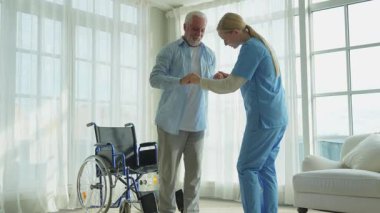 Rehabilitasyon sağlık hizmetleri. Fizyoterapist hemşire tekerlekli sandalyedeki yaşlı adamın ayağa kalkmasına yardım ediyor. Engelli insanlar için sandalyedeki adam hemşire yardımı ile birkaç adım atıyor. Tıbbi bakıcı.