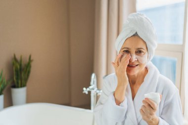 Yetişkin yaşlı bir kadın günlük güzellik rutini yaparak anti-yaş yüz kremi sürüyor. Beyaz maskeli ya da cildinde krem olan yaşlı bir kadın cilt bakımına gidiyor.