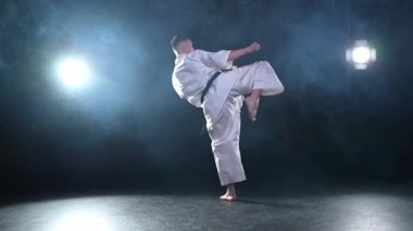 Siyah kuşaklı karate dövüş sanatları için beyaz kimonolu güçlü adam siyah arka plandaki dövüş eğitimine hazırlanıyor. Antrenman hareketleri yumruklar gölge dövüşü alıştırmaları antrenmana hazır.