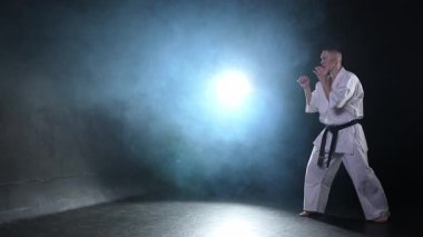Siyah kuşaklı karate dövüş sanatları için beyaz kimonolu güçlü adam siyah arka plandaki dövüş eğitimine hazırlanıyor. Antrenman hareketleri yumruklar gölge dövüşü alıştırmaları antrenmana hazır.