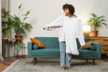 Moda alışverişi. Evde elbise seçen Afrikalı bir kadın. Elbise seçmeye çalışan genç bir kadın. Elbiseleri askıda tutan bir elbise. Kıvırcık esmer tenli, gülümseyen kız elinde askıyla beyaz elbiseyle