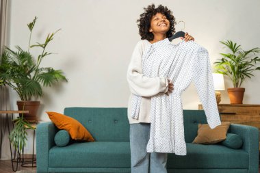 Moda alışverişi. Evde elbise seçen Afrikalı bir kadın. Elbise seçmeye çalışan genç bir kadın. Elbiseleri askıda tutan bir elbise. Kıvırcık esmer tenli, gülümseyen kız elinde askıyla beyaz elbiseyle