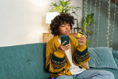 African american woman shopping online holding smartphone paying with gold credit card. Girl sitting at home buying on Internet enter credit card details. Online shopping ecommerce delivery service