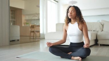 Yoga farkındalığı meditasyonu. Genç sağlıklı Afrikalı kız evde yoga yapıyor. Lotus 'ta oturan kadın yoga minderinde poz veriyor içeride meditasyon yapıyor. Nefes egzersizi yapan bir kız. Evde yoga.