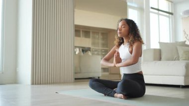 Yoga farkındalığı meditasyonu. Genç sağlıklı Afrikalı kız evde yoga yapıyor. Lotus 'ta oturan kadın yoga minderinde poz veriyor içeride meditasyon yapıyor. Nefes egzersizi yapan bir kız. Evde yoga.