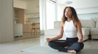 Yoga farkındalığı meditasyonu. Genç sağlıklı Afrikalı kız evde yoga yapıyor. Lotus 'ta oturan kadın yoga minderinde poz veriyor içeride meditasyon yapıyor. Nefes egzersizi yapan bir kız. Evde yoga.