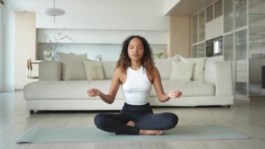 Yoga farkındalığı meditasyonu. Genç sağlıklı Afrikalı kız evde yoga yapıyor. Lotus 'ta oturan kadın yoga minderinde poz veriyor içeride meditasyon yapıyor. Nefes egzersizi yapan bir kız. Evde yoga.