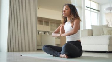 Yoga farkındalığı meditasyonu. Genç sağlıklı Afrikalı kız evde yoga yapıyor. Lotus 'ta oturan kadın yoga minderinde poz veriyor içeride meditasyon yapıyor. Nefes egzersizi yapan bir kız. Evde yoga.