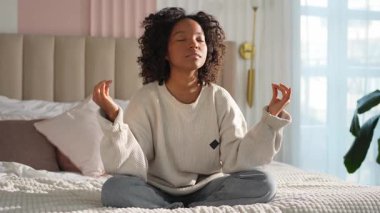 Yoga farkındalığı meditasyonu. Genç sağlıklı Afrikalı kız evde yoga yapıyor. Nilüfer çiçekli bir kadın yatakta oturmuş, meditasyon yapıyor, gülümsüyor, evde dinleniyor. Nefes egzersizi yapan bir kız. Evde yoga.