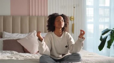 Yoga farkındalığı meditasyonu. Genç sağlıklı Afrikalı kız evde yoga yapıyor. Nilüfer çiçekli bir kadın yatakta oturmuş, meditasyon yapıyor, gülümsüyor, evde dinleniyor. Nefes egzersizi yapan bir kız. Evde yoga.
