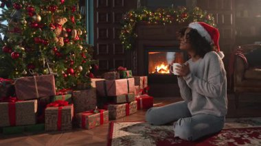 Mutlu noeller. Noel Baba şapkalı Afro-Amerikalı kadın, koyu renkli bir Noel ağacının yanında bir fincan sıcak kahve çayıyla. Oturma odasında Noel ağacı ve şöminesi olan bir kız. Evde Noel arifesi