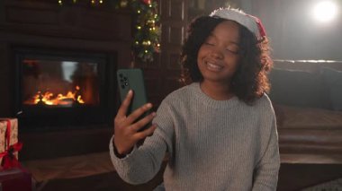 Mutlu noeller. Noel Baba şapkalı Afrikalı kadın akıllı telefondan görüntülü konuşma yapıyor. Sosyal ağda neşeli ruh hali kayıt videosu blogcusu. Noel 'i kutlamak için arkadaşlarıyla görüntülü görüşme yapan bir kız.