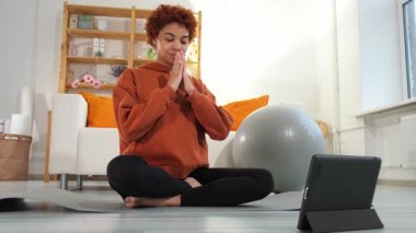 Yoga mindfulness meditation. Young healthy african girl practicing yoga at home. Woman sitting in lotus pose on yoga mat eyes close hands in prayer meditating indoor. Girl doing breathing practice