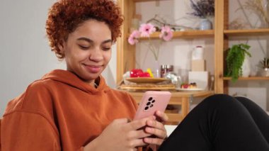 African girl holding smartphone touch screen typing scroll page at home. Woman with cell phone surfing internet using social media apps playing game. Shopping online Internet news cellphone addiction