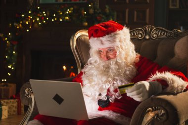 Mutlu noeller. Noel Baba internetten alışveriş yapıyor elinde dizüstü bilgisayar var. Noel hediyeleri için kredi kartıyla şenlikli oturma odasında ödeme yapıyor. Noel Baba Noel arifesinde hediye kutusu alıyor. Kış Tatili Kutlamaları