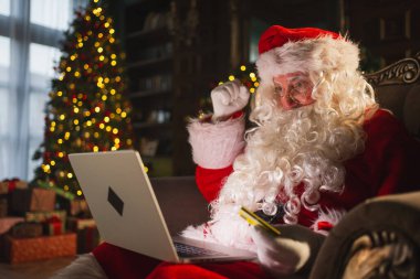 Mutlu noeller. Noel Baba internetten alışveriş yapıyor elinde dizüstü bilgisayar var. Noel hediyeleri için kredi kartıyla şenlikli oturma odasında ödeme yapıyor. Noel Baba Noel arifesinde hediye kutusu alıyor. Kış Tatili Kutlamaları