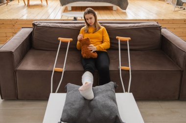 Kanepede oturan kırık bacaklı genç kız. Üzgün küçük çocuk, kırık bacağını iyileştirmek için oyuncak ayısına sarılıyor. Alçı ve koltuk değneği kullanan bir kız. Kaza sonucu oluşan kırık, ayak bandajı.