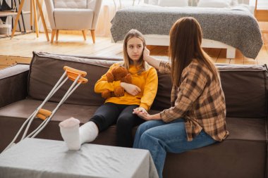 Kırık bacağı alçıda olan bir kız annesiyle evde kanepede oturuyor. Kırık bacağı iyileşen üzgün küçük çocuk oyuncak ayıya sarılıyor. Hızlı rehabilitasyon için koltuk değneklerini kullanan bir kız. Anne destek kızı