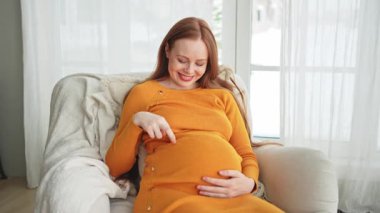 Hamilelik anneliği insanların gelecek beklentisi. Koca göbekli hamile bir kadın evdeki pencerenin yanındaki sandalyeye oturuyor. Karnına sarılan ve hamileliğin tadını çıkaran bir kız. Annelik şefkati ebeveynlik yeni bir hayat