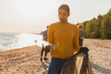 Dünya günü. Gönüllü eylemciler, sahil bölgesindeki çöpleri topluyor. Kadın, okyanus kıyısındaki çöp poşetine plastik şişe koyuyor. Çevre koruma kıyı bölgesi temizliği
