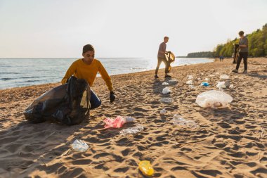 Dünya günü. Gönüllü eylemciler sahil bölgesindeki çöpleri topluyor. Kadınlar ve erkekler okyanus kıyısındaki çöp torbasına plastik çöp koyar. Çevre koruma kıyı bölgesi temizliği