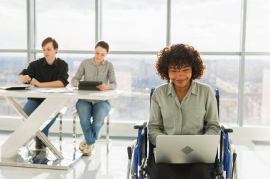 Erişilebilir çevre. Modern ofiste dizüstü bilgisayarla çalışan engelliler için sandalyeli Afrikalı kadın. Birlikte çalışan çok ırklı bir grup insan. Genç profesyonel takım