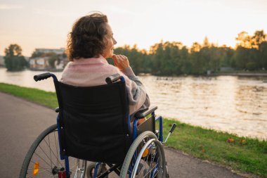 Tekerlekli sandalyede oturan üzgün yaşlı kadın şehir dışında, arka manzaralı bir yolda yürüyor. Engelliler için tekerlekli sandalyedeki yaşlı kadın depresyonda. Yalnız yaşlı hasta kadın tekerlekli sandalyede oturuyor.