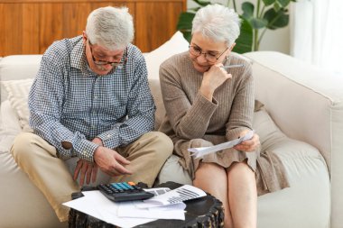 Orta yaşlı yaşlı çift dizüstü bilgisayarları ve kağıt belgeleriyle oturuyor. Yaşlı, olgun bir kadın kağıt faturası okuyor. Ev yöneticiliği yapıyor. Vergileri finanse ediyor.