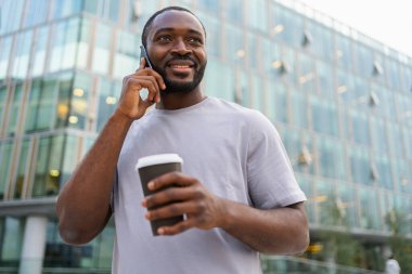 İyi sabahlar. Afrikalı bir adam dışarıda akıllı telefondan konuşmak için kahve içiyor. Elinde cep telefonu bardağı olan bir adam sokakta yürüyor. Cep telefonuyla cevap veren adam cep telefonuyla konuşuyor.