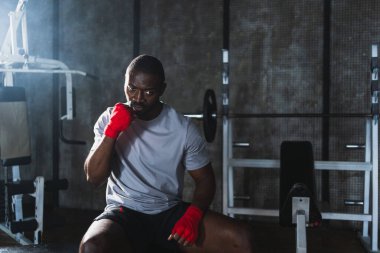 Beden eğitiminde spor. Afrikalı dövüşçü dövüşmeye hazırlanıyor kırmızı boksla yumruk atarken spor sargılarını spor salonunda sarar. Güçlü adam boks antrenmanına hazır yumrukları eğitiyor.