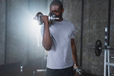 Beden eğitiminde spor. Spor salonunda dambılları toplayan Afrikalı Amerikalı bir vücut geliştirici. Fitness kulübünde halterli kasları çalıştırmak için ağırlık çalışıyorum. Spor yapan bir adam. Sağlıklı yaşam tarzı