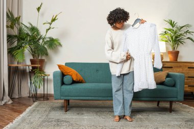 Moda alışverişi. Evde elbise seçen Afrikalı bir kadın. Elbise seçmeye çalışan genç bir kadın. Elbiseleri askıda tutan bir elbise. Kıvırcık esmer tenli, gülümseyen kız elinde askıyla beyaz elbiseyle