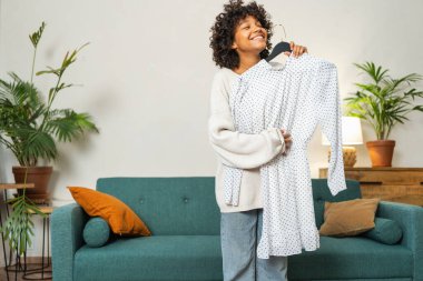 Moda alışverişi. Evde elbise seçen Afrikalı bir kadın. Elbise seçmeye çalışan genç bir kadın. Elbiseleri askıda tutan bir elbise. Kıvırcık esmer tenli, gülümseyen kız elinde askıyla beyaz elbiseyle