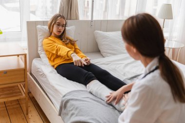 Bacağı kırık bir kız ve klinikte doktor. Travmatolog ortopedist muayenehanede çocuk bacağını muayene ediyor. Kırık ayak bileği yaralanmasından sonra bacağında alçı olan kızı muayene eden doktor.