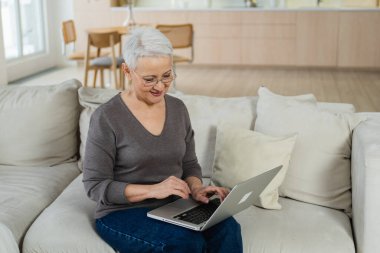 Evde dizüstü bilgisayar kullanan kendinden emin, mutlu, orta yaşlı bir kadın. Yaşlı, şık 60 'lı yılların büyükannesi kanepeye oturmuş bilgisayar ekranına bakıyor daktilo yazarak sohbet ediyor e-posta yazıyor.