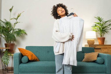 Moda alışverişi. Evde elbise seçen Afrikalı bir kadın. Elbise seçmeye çalışan genç bir kadın. Elbiseleri askıda tutan bir elbise. Kıvırcık esmer tenli, gülümseyen kız elinde askıyla beyaz elbiseyle