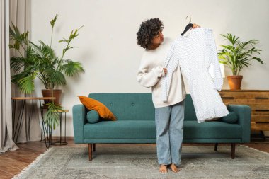 Moda alışverişi. Evde elbise seçen Afrikalı bir kadın. Elbise seçmeye çalışan genç bir kadın. Elbiseleri askıda tutan bir elbise. Kıvırcık esmer tenli, gülümseyen kız elinde askıyla beyaz elbiseyle