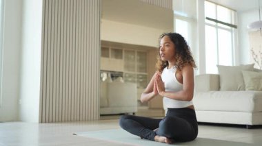 Yoga farkındalığı meditasyonu. Genç sağlıklı Afrikalı kız evde yoga yapıyor. Lotus 'ta oturan kadın yoga minderinde poz veriyor içeride meditasyon yapıyor. Nefes egzersizi yapan bir kız. Evde yoga.
