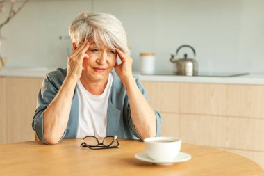 Baş ağrısı. Mutsuz, orta yaşlı, baş ağrısı çeken, evde şakak ovalayan yaşlı bir kadın. Yaşlı, olgun, yaşlı bir büyükanne tapınaklara dokunuyor. Stresli bir kadın. Başında ağrı hissediyor.