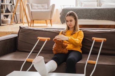 Kanepede oturan kırık bacaklı genç kız. Üzgün küçük çocuk, kırık bacağını iyileştirmek için oyuncak ayısına sarılıyor. Alçı ve koltuk değneği kullanan bir kız. Kaza sonucu oluşan kırık, ayak bandajı.