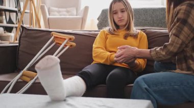 Kırık bacağı alçıda olan bir kız evde kanepede annesiyle oturuyor. Kırık bacağı iyileşen üzgün genç bir çocuk. Hızlı rehabilitasyon için koltuk değneklerini kullanan bir kız. Anne destek kızı