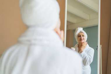 Yetişkin, yaşlı bir kadın aynaya bakıyor ve günlük güzellik kremi sürüyor. Cildinde beyaz maske kremi olan yaşlı bir kadın cilt bakımı görüyor.