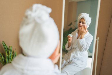 Yetişkin, yaşlı bir kadın aynaya bakıyor ve günlük güzellik kremi sürüyor. Cildinde beyaz maske kremi olan yaşlı bir kadın cilt bakımı görüyor.