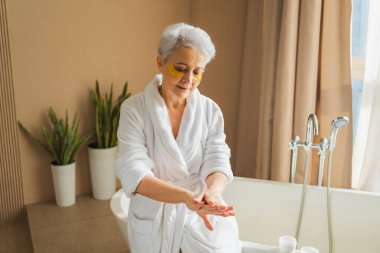 Olgun, yaşlı bir kadın ellerinde nemlendirici vücut losyonu sürüyor. Son sınıf öğrencisi kadın banyoda günlük cilt bakımını yaparken anti-yaş kremi sürüyor. Güzellik tedavisi vücut bakımını kapsar.