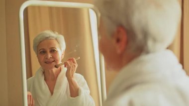 Yumuşak, pürüzsüz bir teni olan, yanağında fırçayla aynaya bakıp günlük güzellik makyajı yapan olgun, yaşlı bir kadın. Kıdemli kadın yüzüne allık sürüyor. Yaş karşıtı makyaj