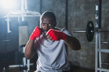 Beden eğitiminde spor. Afrikalı dövüşçü dövüşmeye hazırlanıyor kırmızı boksla yumruk atarken spor sargılarını spor salonunda sarar. Güçlü adam boks antrenmanına hazır yumrukları eğitiyor.